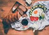 A rustic wooden table is set with a variety of breakfast items. A loaf of brown bread is partially sliced on a cutting board with a pat of butter nearby. Next to it, there's a plate featuring a fried egg surrounded by sliced cucumbers, tomato slices, and greens. A cup of tea sits adjacent to a white teapot, all set on a delicate floral cloth.