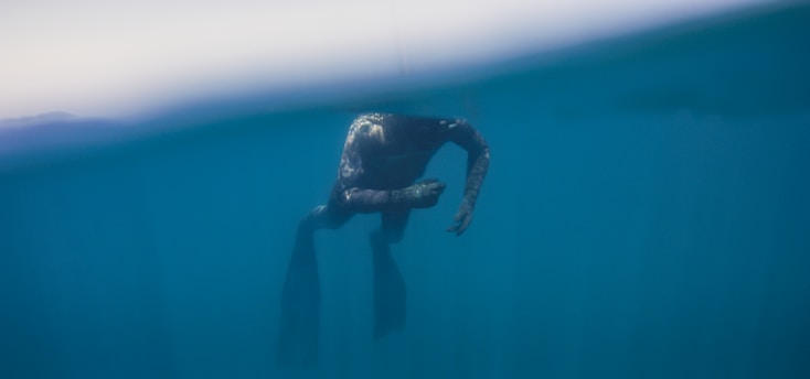 A diver is submerged in deep blue water, with most of the figure visible beneath the surface. The light creates a gradient effect, with lighter hues near the surface transitioning to darker shades below.