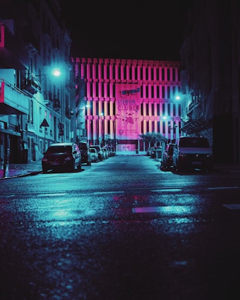 A city street scene at night, illuminated by vibrant neon pink and blue lights. The street is lined with parked cars on both sides, leading towards a building with a brightly lit facade. The building features a large banner with the words 'LISBOA GIN&Aacute;SIO CLUBE'. The wet pavement reflects the colorful lights, adding to the atmospheric effect.