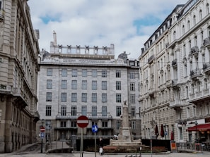 A historic European cityscape featuring an ornate building with 'Österreichische Postsparkasse' written at the top. Surrounded by classical architecture, the area includes a notable stone monument in the foreground. The scene is set under a partly cloudy sky, with some street signs and a few pedestrians visible.