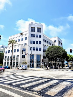A modern, multi-story office building with white exterior walls and large rectangular windows. The structure is situated at a busy intersection with crosswalks. Several people are walking on the sidewalks, and there are cars on the street, including a postal vehicle. Palm trees and other lush greenery line the street, and the sky is clear and bright blue.