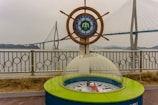 A maritime university emblem is set within a ship's wheel, positioned beside a decorative compass enclosed in glass. In the background is a large suspension bridge spanning across water, with overcast skies adding a muted tone to the scene.