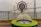 A maritime university emblem is set within a ship's wheel, positioned beside a decorative compass enclosed in glass. In the background is a large suspension bridge spanning across water, with overcast skies adding a muted tone to the scene.