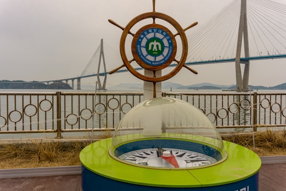 A maritime university emblem is set within a ship's wheel, positioned beside a decorative compass enclosed in glass. In the background is a large suspension bridge spanning across water, with overcast skies adding a muted tone to the scene.