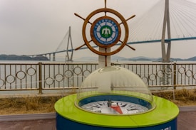 A maritime university emblem is set within a ship's wheel, positioned beside a decorative compass enclosed in glass. In the background is a large suspension bridge spanning across water, with overcast skies adding a muted tone to the scene.
