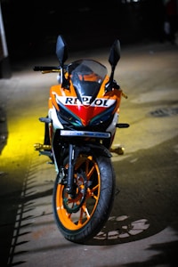 A motorcycle with a vibrant orange and white color scheme parked on a dimly lit street. The brand name 'Repsol' is prominently displayed on the front. The bike features black side mirrors and orange wheels. The lighting casts dramatic shadows around the vehicle.