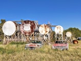 A series of large, rusted cylindrical tanks are arranged on wooden supports. In front of the tanks, two vintage cars with weathered exteriors are parked in an overgrown grassy field. The tanks, bearing the 'Phillips 66' logo, suggest a bygone era of fuel storage or distribution.
