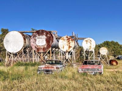 A series of large, rusted cylindrical tanks are arranged on wooden supports. In front of the tanks, two vintage cars with weathered exteriors are parked in an overgrown grassy field. The tanks, bearing the 'Phillips 66' logo, suggest a bygone era of fuel storage or distribution.