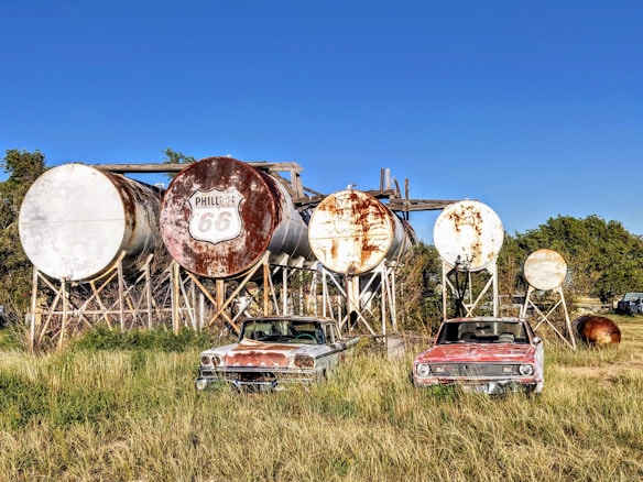 A series of large, rusted cylindrical tanks are arranged on wooden supports. In front of the tanks, two vintage cars with weathered exteriors are parked in an overgrown grassy field. The tanks, bearing the 'Phillips 66' logo, suggest a bygone era of fuel storage or distribution.