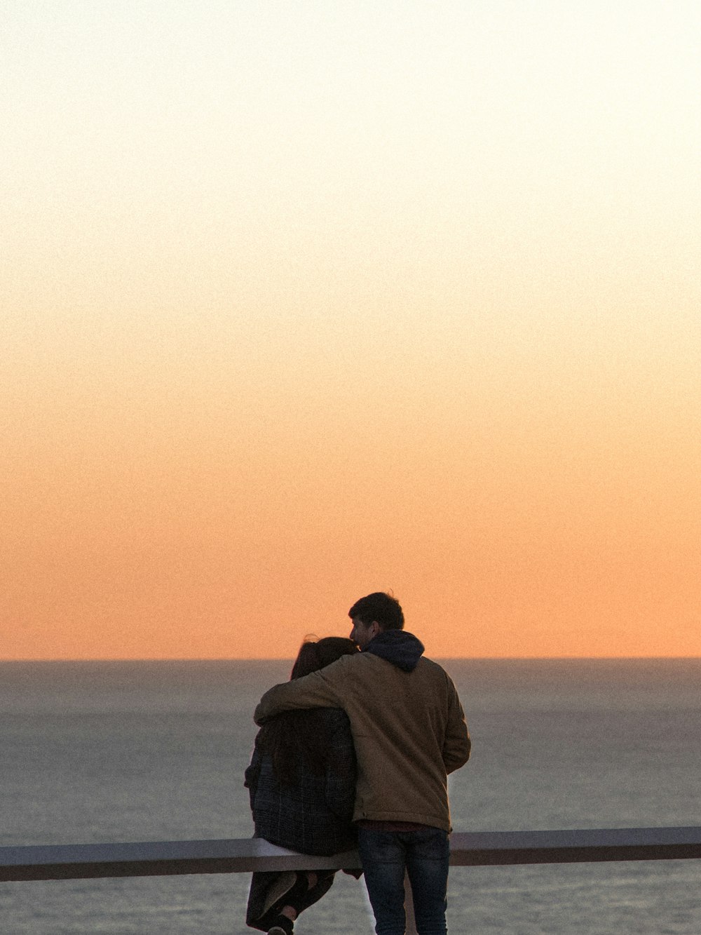 man in brown jacket and black knit cap sitting on brown rock during sunset