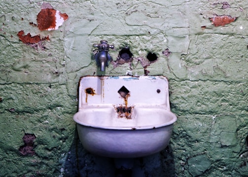 An old, rusty sink is mounted on a worn and peeling green wall. The surface of the wall shows signs of decay and neglect, with exposed patches and holes. The faucet above the sink is metal and similarly corroded, contributing to the overall sense of age and disrepair.