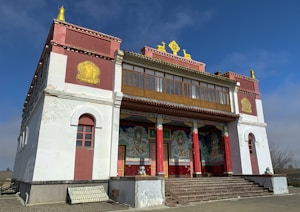A traditional Buddhist temple with ornate decorations and vibrant colors. The building has a rectangular structure with a white and red facade, golden ornaments, and intricate paintings near the entrance. There are large steps leading up to the entrance, which is supported by red columns.