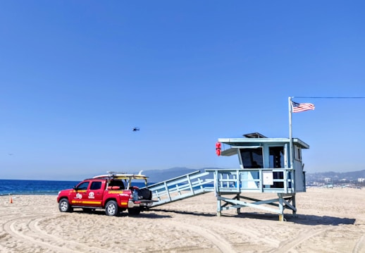 A lifeguard station on a sandy beach with a red lifeguard truck parked nearby. There is an American flag flying on a pole attached to the station. The truck is marked with lifeguard insignia and has a surfboard on its roof rack. In the background, the ocean and distant hills are visible under a clear blue sky. A small aircraft or drone can be seen in the sky.