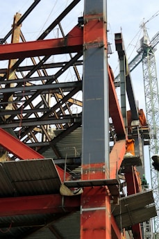 A construction site with a steel framework structure and a tall crane. The framework consists of red and grey beams, partially completed with some sections showing metal sheeting. The crane is green, towering in the background, indicating active construction.