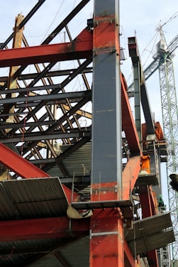 A construction site with a steel framework structure and a tall crane. The framework consists of red and grey beams, partially completed with some sections showing metal sheeting. The crane is green, towering in the background, indicating active construction.