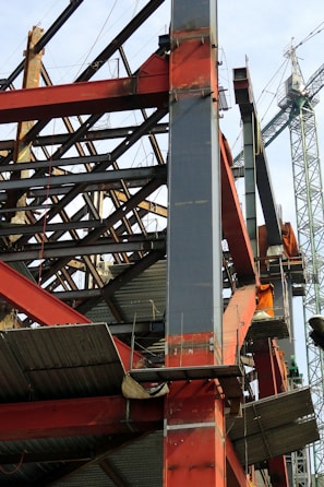 A construction site with a steel framework structure and a tall crane. The framework consists of red and grey beams, partially completed with some sections showing metal sheeting. The crane is green, towering in the background, indicating active construction.