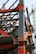 A construction site with a steel framework structure and a tall crane. The framework consists of red and grey beams, partially completed with some sections showing metal sheeting. The crane is green, towering in the background, indicating active construction.