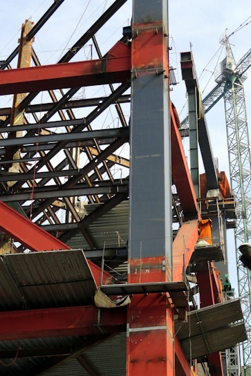 A construction site with a steel framework structure and a tall crane. The framework consists of red and grey beams, partially completed with some sections showing metal sheeting. The crane is green, towering in the background, indicating active construction.