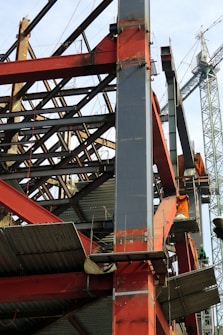 A construction site with a steel framework structure and a tall crane. The framework consists of red and grey beams, partially completed with some sections showing metal sheeting. The crane is green, towering in the background, indicating active construction.