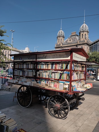An outdoor book cart filled with a variety of books is displayed on a paved street. The cart has large wooden wheels and is positioned in front of an ornate, historic building with domed roofs and intricate architecture. The area is well-lit with clear blue skies above, and there is foliage from nearby trees partially visible. This scene appears to be in an urban setting, with some cars and people in the background.