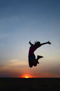 A sunset silhouette of a person jumping joyfully on a hilltop.