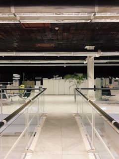 A modern office space featuring a pathway with glass railings on either side. The floor is made of white marble tiles, leading towards a workspace with office chairs, filing cabinets, and a visible potted plant. The ceiling has a grid-like pattern with exposed ducts and lighting fixtures.