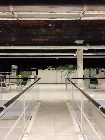 A modern office space featuring a pathway with glass railings on either side. The floor is made of white marble tiles, leading towards a workspace with office chairs, filing cabinets, and a visible potted plant. The ceiling has a grid-like pattern with exposed ducts and lighting fixtures.