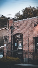 A rustic brick cafe building with a black framed glass door, flanked by small trees wrapped in lights. There are vintage-style black lamps on the walls and a sign on the door with text about Sausalito Equator. In the background, greenery and power lines are visible.