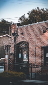 A rustic brick cafe building with a black framed glass door, flanked by small trees wrapped in lights. There are vintage-style black lamps on the walls and a sign on the door with text about Sausalito Equator. In the background, greenery and power lines are visible.
