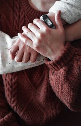 A close-up of two people wearing textured knit sweaters in earthy tones, one arm wrapped around the other. One person is wearing a speckled cream sweater, while the other wears a rich, brown cable-knit sweater. The person in the cream sweater has a smart fitness tracker on their wrist.