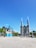A large, gothic-style cathedral with twin spires dominates the background. In the foreground, there is a massive black spider sculpture. Tourists are walking and gathering around the area, enjoying a bright, clear day. The surroundings are urban with trees and some modern buildings.