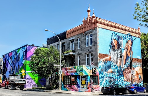 A vibrant urban scene featuring two buildings with colorful murals. The building on the right has a large mural of two figures with blue checkered patterns. The left building displays an abstract geometric design with bright colors. A truck is parked on the street alongside cars. Trees and street signs are visible, giving a lively atmosphere.