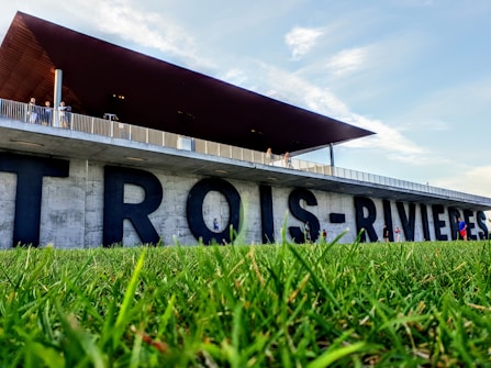 Large, bold letters spelling 'TROIS-RIVIERES' are affixed to a concrete structure. Above, a modern building with a wooden overhang extends outwards, with people walking along the balcony. The foreground features vibrant green grass and a bright blue sky is visible.