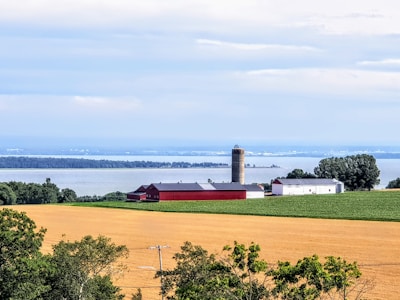 A tranquil rural landscape featuring a farm with red and white buildings, a silo, and fields of crops bordered by trees. In the background, a large body of water stretches across the horizon under a cloudy blue sky.