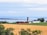 A tranquil rural landscape featuring a farm with red and white buildings, a silo, and fields of crops bordered by trees. In the background, a large body of water stretches across the horizon under a cloudy blue sky.