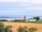 A tranquil rural landscape featuring a farm with red and white buildings, a silo, and fields of crops bordered by trees. In the background, a large body of water stretches across the horizon under a cloudy blue sky.