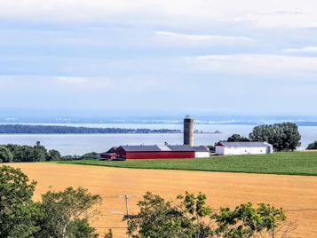 A tranquil rural landscape featuring a farm with red and white buildings, a silo, and fields of crops bordered by trees. In the background, a large body of water stretches across the horizon under a cloudy blue sky.