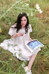 A woman with long dark hair sits in tall grass, wearing a white dress with delicate patterns. She holds a glass of red liquid in one hand and a book open on her lap with an image of clouds. The setting is outdoors with greenery and flowers surrounding her.