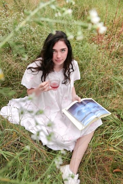 A woman with long dark hair sits in tall grass, wearing a white dress with delicate patterns. She holds a glass of red liquid in one hand and a book open on her lap with an image of clouds. The setting is outdoors with greenery and flowers surrounding her.