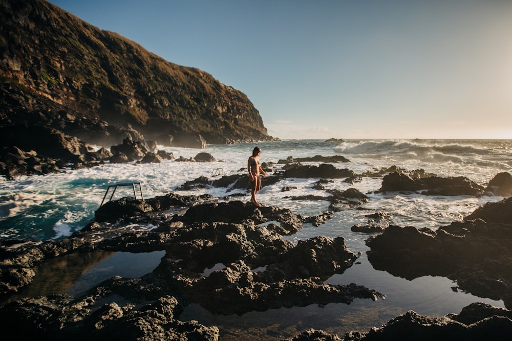 woman in white bikini standing on rocky shore during daytime