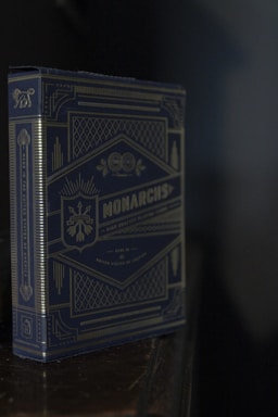 A deck of playing cards in a blue and gold ornate box with intricate geometric and botanical designs. The box features the word 'Monarchs' prominently in the center with a stylish, regal theme.