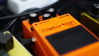 An orange guitar effects pedal labeled 'Distortion DS-1' with black knobs for adjusting output and input. Surrounded by cables and other pedals, the focus is on the details of the device, showing the connection jack and part of the pedal surface.