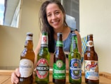 A woman smiling and sitting at a table with five different beer bottles arranged in front of her. The bottles include brands like Hoegaarden, Beck's, Eisenbahn, Heineken, and another with colorful labeling. The setting appears casual and relaxed, with natural light coming through a window.