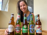 A woman smiling and sitting at a table with five different beer bottles arranged in front of her. The bottles include brands like Hoegaarden, Beck's, Eisenbahn, Heineken, and another with colorful labeling. The setting appears casual and relaxed, with natural light coming through a window.