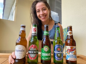 A woman smiling and sitting at a table with five different beer bottles arranged in front of her. The bottles include brands like Hoegaarden, Beck's, Eisenbahn, Heineken, and another with colorful labeling. The setting appears casual and relaxed, with natural light coming through a window.