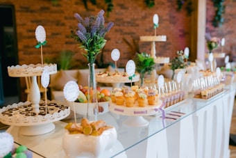 An elegantly arranged dessert table displaying a variety of sweets, including cupcakes, cookies, and candies. The setup is decorated with small bouquets of purple and green flowers and set against a backdrop of a brick wall. The table features white cake stands and decorative cards with green ribbons as labels.