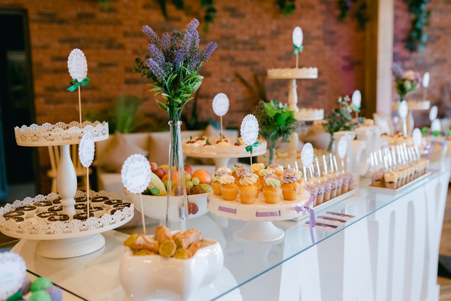 An elegantly arranged dessert table displaying a variety of sweets, including cupcakes, cookies, and candies. The setup is decorated with small bouquets of purple and green flowers and set against a backdrop of a brick wall. The table features white cake stands and decorative cards with green ribbons as labels.