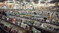 A large record store filled with rows of vinyl records and CDs. Shelves display various album covers in diverse genres, and the walls are covered with posters. Several people browse through the collection.