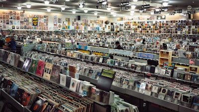 A large record store filled with rows of vinyl records and CDs. Shelves display various album covers in diverse genres, and the walls are covered with posters. Several people browse through the collection.