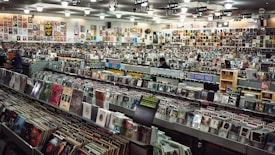 A large record store filled with rows of vinyl records and CDs. Shelves display various album covers in diverse genres, and the walls are covered with posters. Several people browse through the collection.